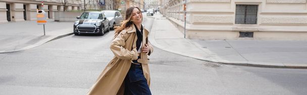 cheerful young woman in beige trench coat walking on european street, banner - Photo, Image