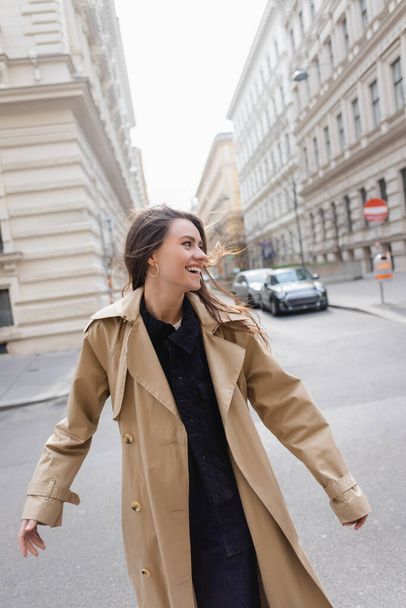 joyful young woman in beige trench coat walking on european street - Foto, afbeelding