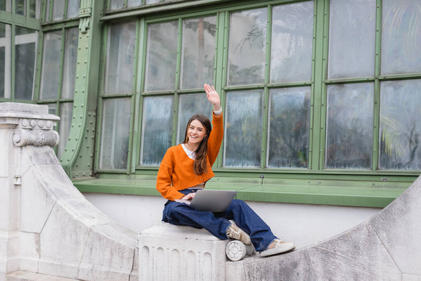 happy young woman waving hand while using laptop on rooftop of building in vienna  - Foto, immagini