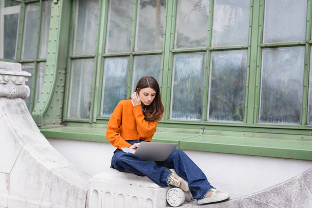 young woman in jeans and orange cardigan using laptop on rooftop of building - Φωτογραφία, εικόνα