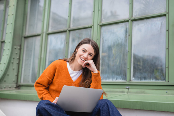 happy young woman in jeans and orange cardigan using laptop on rooftop of building - Photo, image