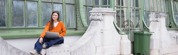 smiling young freelancer in jeans and orange cardigan sitting with laptop on rooftop of building, banner - Foto, Imagem