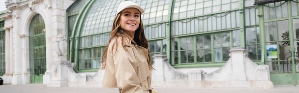 cheerful young woman in trench coat and baseball cap standing on rooftop of european building, banner - Fotó, kép