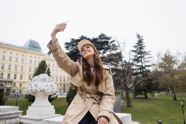 happy young woman in stylish trench coat and baseball cap taking selfie in european city  - Foto, Imagen