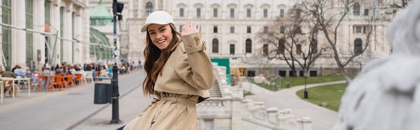 happy young woman in stylish trench coat and baseball cap waving hand outside, banner - Valokuva, kuva