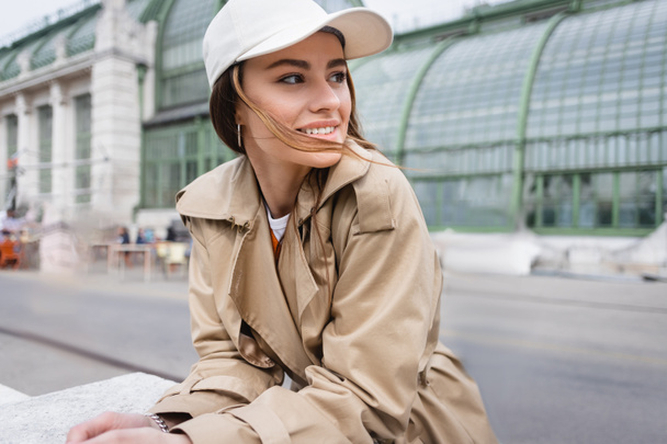 happy young woman in beige trench coat and baseball cap looking away - Photo, Image