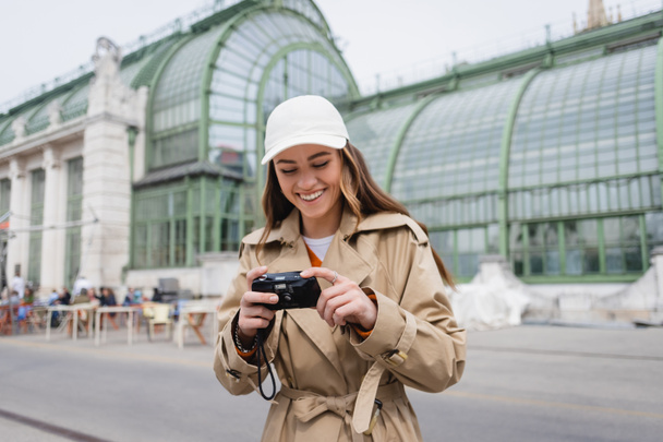 happy young woman in beige trench coat and baseball cap holding vintage camera  - Фото, изображение