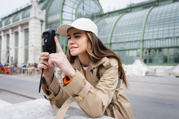 young woman in beige trench coat and baseball cap taking photo on vintage camera  - Photo, Image