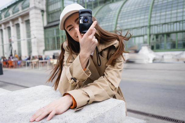 young woman in trench coat and baseball cap taking photo on vintage camera  - Photo, Image