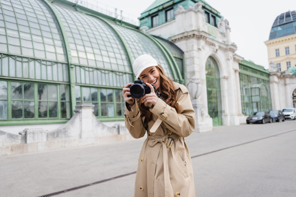 cheerful photographer in trench coat and baseball cap taking photo on digital camera in european city - 写真・画像