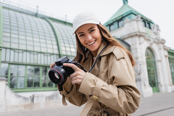 cheerful woman in trench coat and baseball cap holding digital camera in european city - Photo, Image