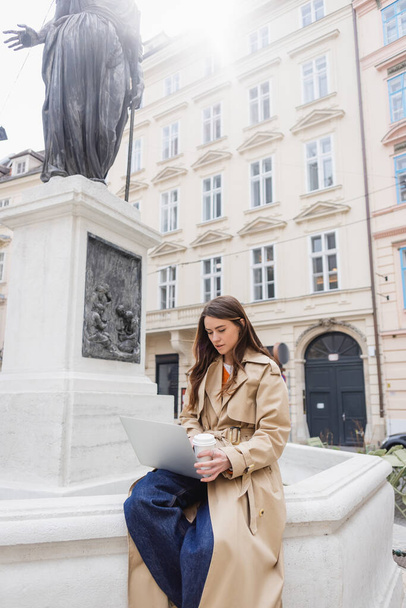 young woman in stylish trench coat looking at laptop and holding paper cup in europe  - Foto, Imagem