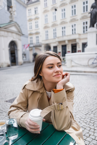 young woman in trendy trench coat holding coffee to go on cafe terrace  - Photo, Image