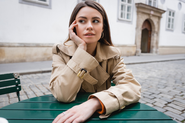 thoughtful young woman in trendy trench coat sitting in summer terrace  - 写真・画像