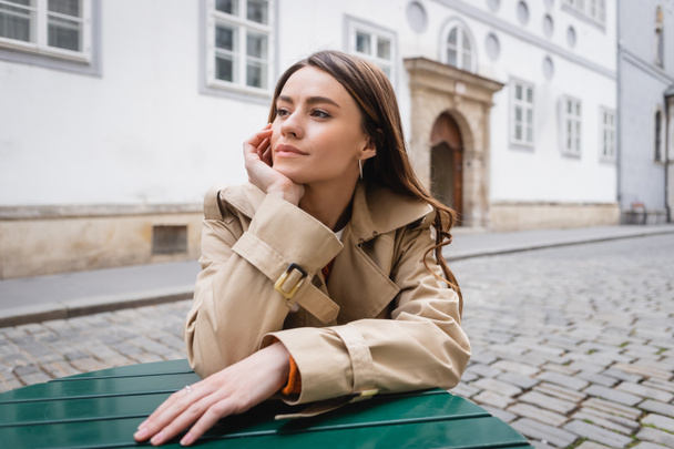 dreamy young woman in trendy trench coat sitting in summer terrace  - Фото, изображение