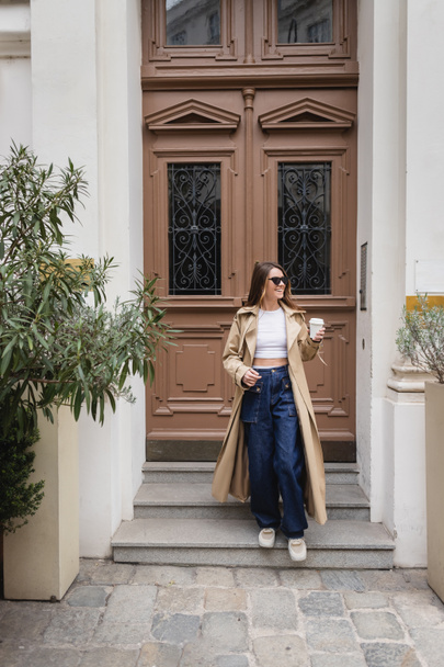 full length of joyful young woman in trendy sunglasses and trench coat holding paper cup near entrance door  - Photo, Image