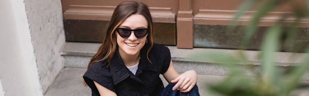 vista ad alto angolo di felice giovane donna in occhiali da sole alla moda seduta vicino alla porta d'ingresso, banner - Foto, immagini
