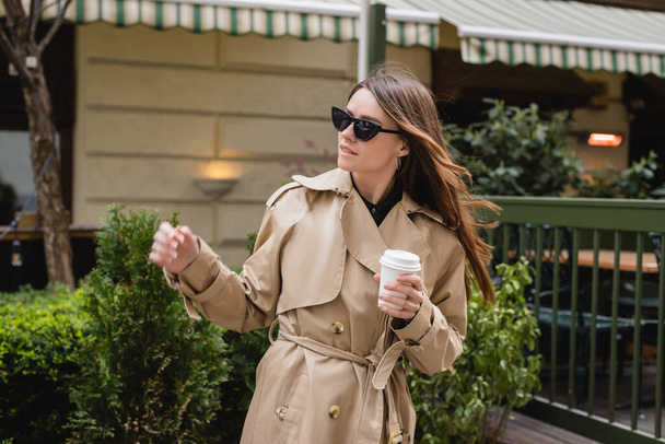 young woman in trendy sunglasses and trench coat holding paper cup  - Photo, Image