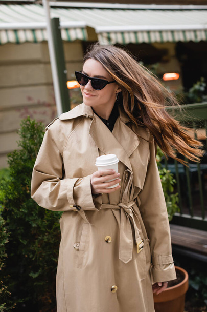 happy young woman in stylish sunglasses and trench coat holding paper cup  - Foto, Bild