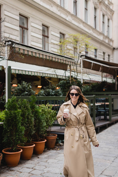 smiling young woman in stylish sunglasses and trench coat holding paper cup outside  - Foto, afbeelding