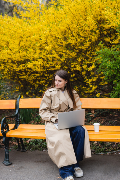 young woman in beige trench coat sitting on bench with laptop near paper cup - Photo, Image