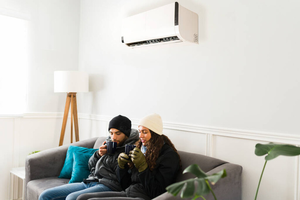 Young couple with the air conditioner unit on drinking hot tea and wearing winter clothes because of the cold air  - Foto, Imagen