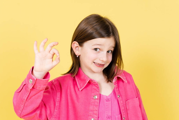 Positive kleine Mädchen machen ok, gut oder fein Zeichen mit den Fingern und Blick auf Kamera isoliert auf gelbem Hintergrund - Foto, Bild