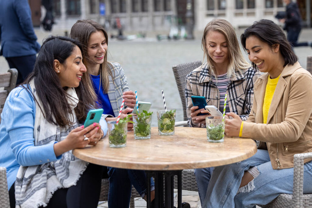 Diverso grupo de jóvenes empresarias discutiendo algo con teléfonos inteligentes en un patio de bar después del trabajo.  - Foto, imagen
