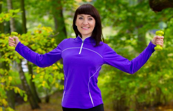 Brunette during fitness exercise with dumbbells in autumn park - Фото, зображення