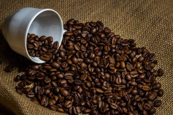 The coffee beans spilled out of the coffee cup. Roasted coffee beans on burlap. - Photo, image