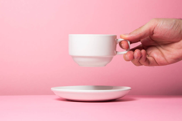 A man's hand holds a white cup on a pink background. Lift the cup from the saucer - 写真・画像