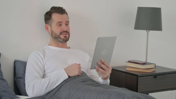 男はベッドでタブレット上でビデオ通話を行う - 写真・画像