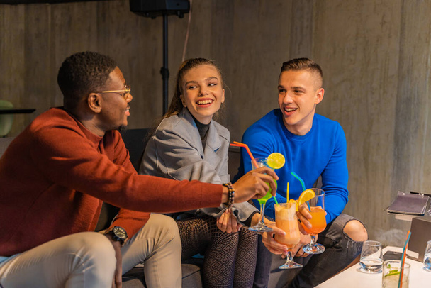 Drei multiethnische Freunde lächeln, während sie zusammen sitzen und mit ihren Getränken im Café jubeln - Foto, Bild