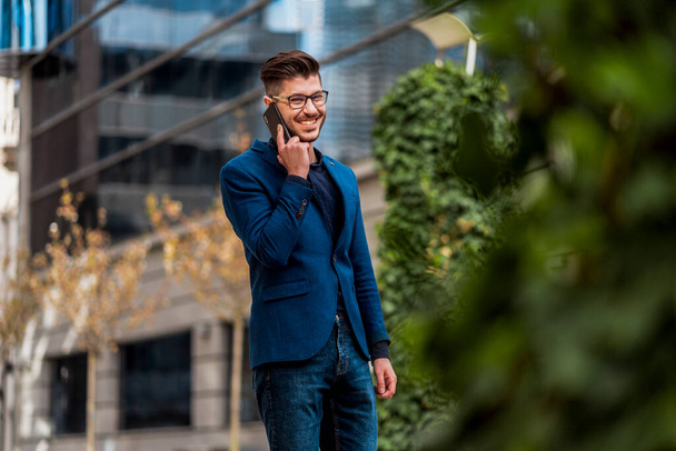 Homme d'affaires urbain en manteau bleu marchant dans la ville et parlant sur téléphone portable - Photo, image