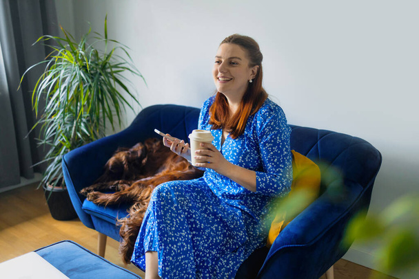 Nuori eurooppalainen hymyilevä 30-vuotias nainen, joka käyttää matkapuhelinta sosiaalisessa mediassa ja nauttii kahvin juomisesta kotona. - Valokuva, kuva
