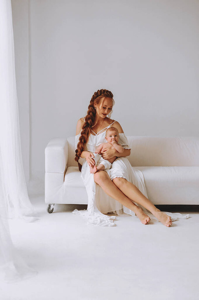 Portrait of redhead woman with long braidhair in white summer dress sitting on white sofa and holding her baby boy. The concept of happy motherhood  - Photo, Image