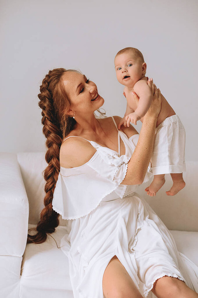 Beyaz yazlık elbiseli, uzun saçlı kızıl saçlı kadın beyaz koltukta oturuyor ve bebeğini elleriyle tutuyor ve gülümsüyor. Mutlu annelik kavramı.  - Fotoğraf, Görsel