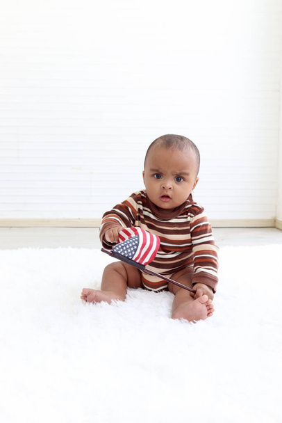 Afro-Amerikaanse baby zittend op de vloer in een overdekte woonkamer, klein kind dat de Amerikaanse vlag vasthoudt en zwaait op 4 juli om de nationale Onafhankelijkheidsdag te vieren, Flag Day concept. - Foto, afbeelding