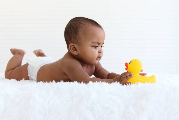 Un bebé afroamericano de seis meses gateando jugando con un juguete de pato amarillo en una alfombra blanca esponjosa, un niño adorable y adorable niña feliz sonriente acostado en la cama en el dormitorio, la infancia y el concepto de cuidado del bebé.. - Foto, Imagen