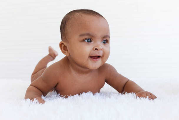 Portrait of a six month crawling African American baby on fluffy white rug, happy smiling adorable sweet little girl kid lying on bed in bedroom, childhood and baby care concept - Photo, Image