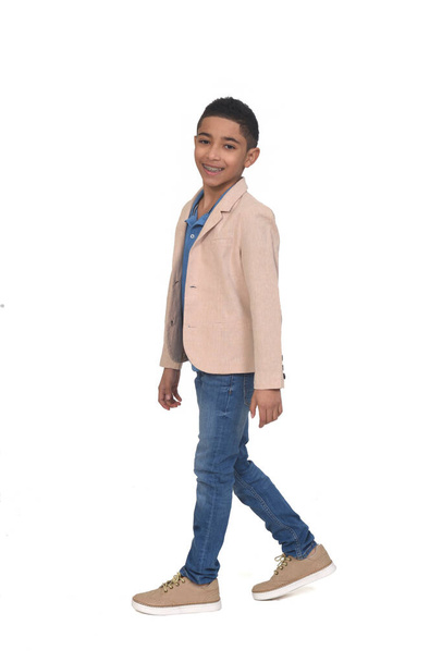 vista laterale del ragazzo con jeans e blazer che cammina e guarda la fotocamera su sfondo bianco - Foto, immagini