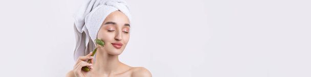 Beautiful girl with thick eyebrows and perfect skin at white background, towel on head, beauty photo. Holds a cosmetic roller. - Φωτογραφία, εικόνα