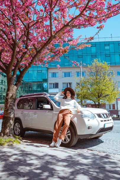 stylish smiling beautiful woman standing near car blooming sakura tree on background spring is coming - Zdjęcie, obraz