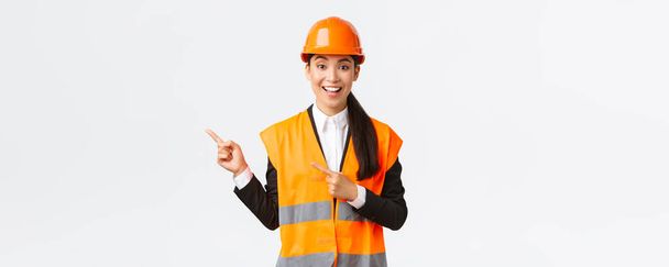 Épület, építőipar és ipari koncepció. Mosolygó női mérnök sisak és reflektív ruházat mutatja az utat, rámutatva a bal felső sarokban, beszél építési projekt, fehér háttér - Fotó, kép
