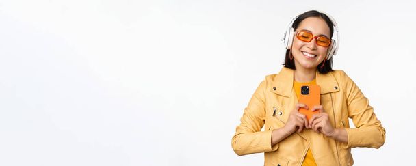 Technology concept. Stylish asian girl in headphones, holding smartphone, dancing and singing, listening music, standing over white background - Fotó, kép