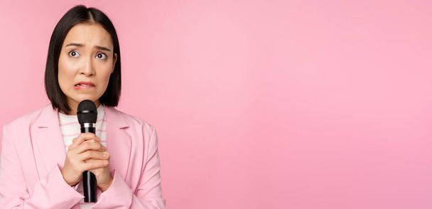 Anxious asian lady in suit, talking in public, giving speech with microphone on conference, looking scared, standing over pink background - Photo, Image