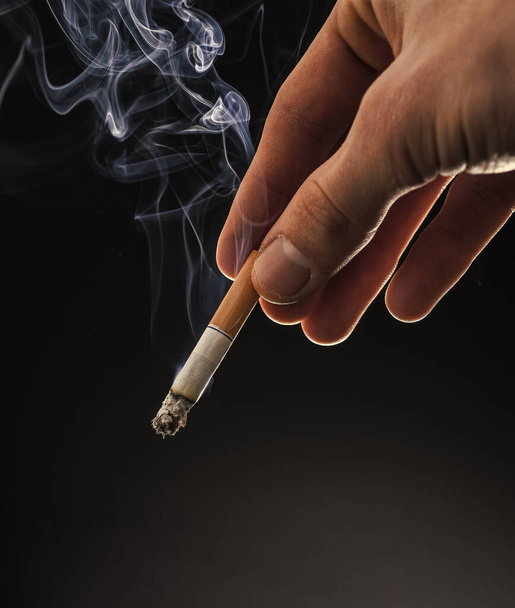 Male hand hold cigarette butt with smoke swirls dark background, copy space, nicotine addiction - Foto, afbeelding