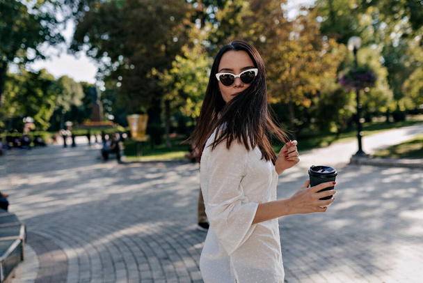 Portret stylu życia stylowej modnej dziewczyny w okularach przeciwsłonecznych nosi elegancką białą bluzkę odwróć się przed kamerą. Zewnętrzne zdjęcie spacerujące w parku w ciepły słoneczny dzień - Zdjęcie, obraz