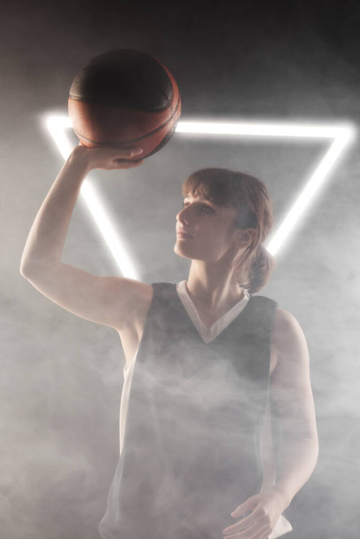 Beautiful girl with basketball. Side lit studio portrait against fog background with white neon lights. - 写真・画像
