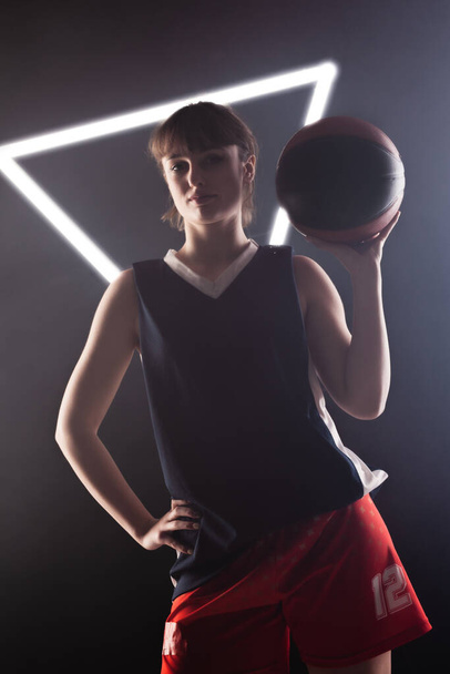 Beautiful girl with basketball. Side lit studio portrait against fog background with white neon lights. - Photo, image